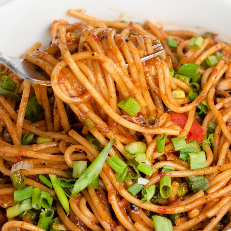 Close-up of Indian noodles with green onions and red peppers, being lifted by a fork.
